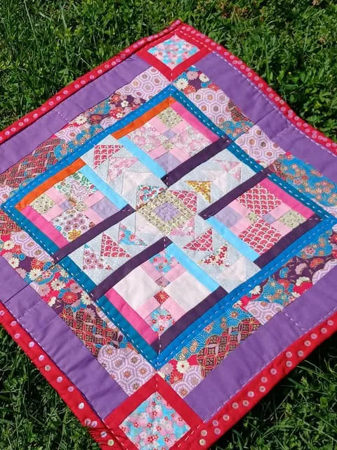 Petit quilt en patchwork rose et violet, posé sur l’herbe
