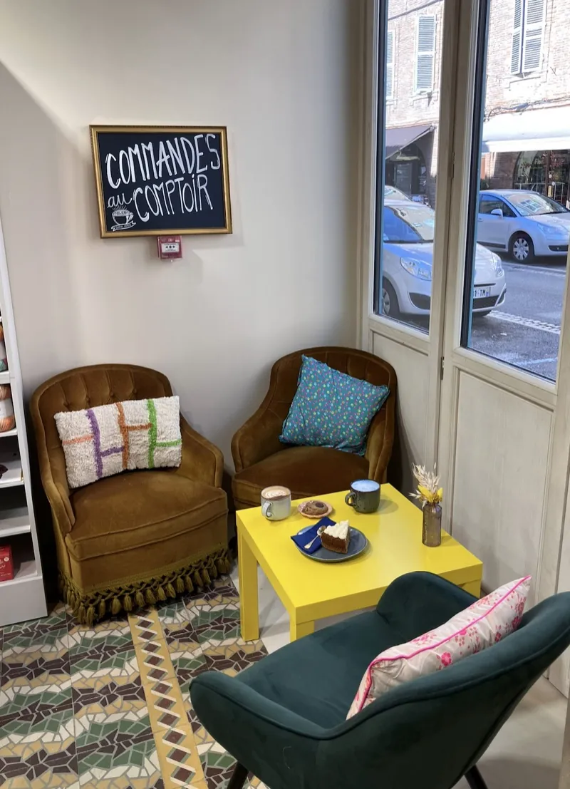 Le coin fenêtre : deux fauteuils en velours, un fauteuil vert et une table jaune avec latte et part de gâteau, sous l’ardoise « Commandes au comptoir »