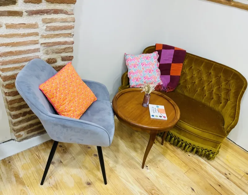 Le coin salon contre le mur en briques : un canapé en velours, un fauteuil gris et une table ronde avec un livre de points de tricot